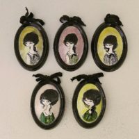 Lot de cinq médaillons en céramique datant des années 50-60, représentant des demoiselles au style et coiffures de cette époque. Chaque médaillons possèdent un noeud en velours noir et une accroche sur l'arrière pour les suspendre au mur. Elégants et romantiques. Signature sur l'arrière. Une micro égrenure sur deux médaillons. Très bon état. Dimensions : 12 x 8,5 cm