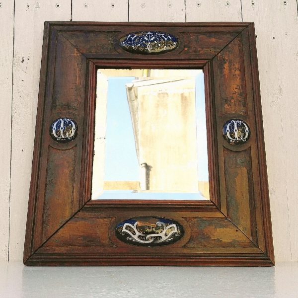 Ancien miroir en bois serti de cabochons émaillés en relief, datant de 1880. Les cabochons de droite et gauche sont monogrammés.  Miroir intérieur épais et biseauté. Des sauts d'émail sur les cabochons haut, bas et de droite. Un point d'accroche sur l'arrière. Bon état général. Dimensions totales : 35 x 39,5 cm Dimensions miroir : 17 x 21 cm