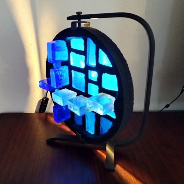 Lampe d'ambiance par Jacques Avoinet, pivotante. Composée d'une structure en fer forgé noir, le diffuseur de lumière est en béton serti de morceaux de verre de couleur bleu. Très bon état. Très lourde.