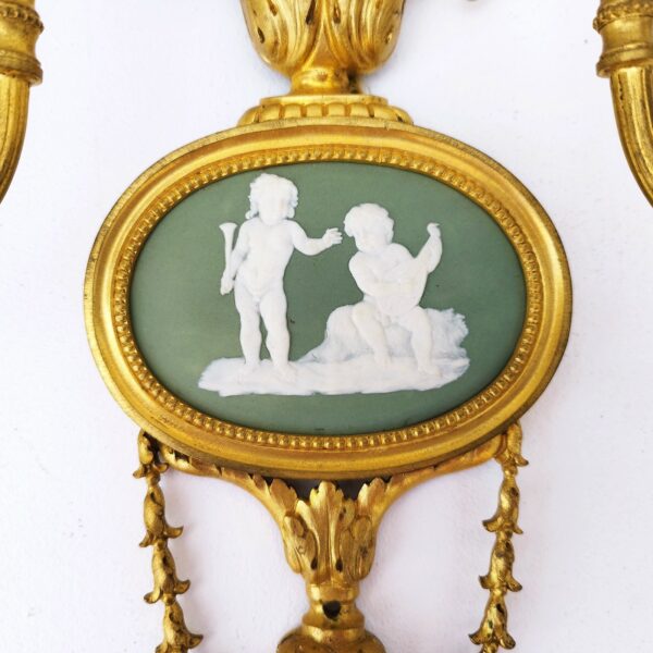 Paire d'appliques en bronze doré de style Louis XVI, médaillon wedgwood au centre au décor d'angelots musiciens. Un noeud ruban sur le haut. Une applique en cassée ( à faire restaurer), un trou de perçage sous le médaillon a été fait certainement pour la fixer solidement au mur. Dans leur jus. Hauteur : 41,5 cm Largeur : 29 cm Profondeur : 11, 5 cm
