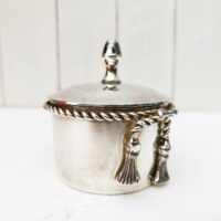 Boite à bijoux en métal argenté de la marque Lancel. Frise torsadée cordelette finissant par deux pompons. Signé Lancel sur le dessous. possède sa boite d'origine. Très bon état Hauteur avec couvercle : 10 cm Diamètre : 7,5 cm