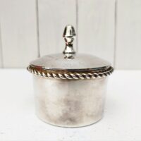 Boite à bijoux en métal argenté de la marque Lancel. Frise torsadée cordelette finissant par deux pompons. Signé Lancel sur le dessous. possède sa boite d'origine. Très bon état Hauteur avec couvercle : 10 cm Diamètre : 7,5 cm