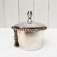 Boite à bijoux en métal argenté de la marque Lancel. Frise torsadée cordelette finissant par deux pompons. Signé Lancel sur le dessous. possède sa boite d'origine. Très bon état Hauteur avec couvercle : 10 cm Diamètre : 7,5 cm