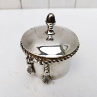 Boite à bijoux en métal argenté de la marque Lancel. Frise torsadée cordelette finissant par deux pompons. Signé Lancel sur le dessous. possède sa boite d'origine. Très bon état Hauteur avec couvercle : 10 cm Diamètre : 7,5 cm