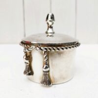 Boite à bijoux en métal argenté de la marque Lancel. Frise torsadée cordelette finissant par deux pompons. Signé Lancel sur le dessous. possède sa boite d'origine. Très bon état Hauteur avec couvercle : 10 cm Diamètre : 7,5 cm
