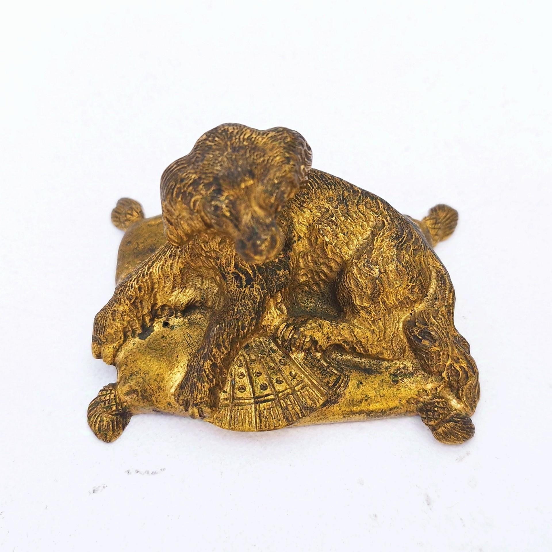 Petit bronze doré représentant un chien assis sur un coussin. quatre pieds représentant les pompons du coussin. Dorure usée par endroit. Bon état général. Dimensions :  7,5 x 5 x 5 cm