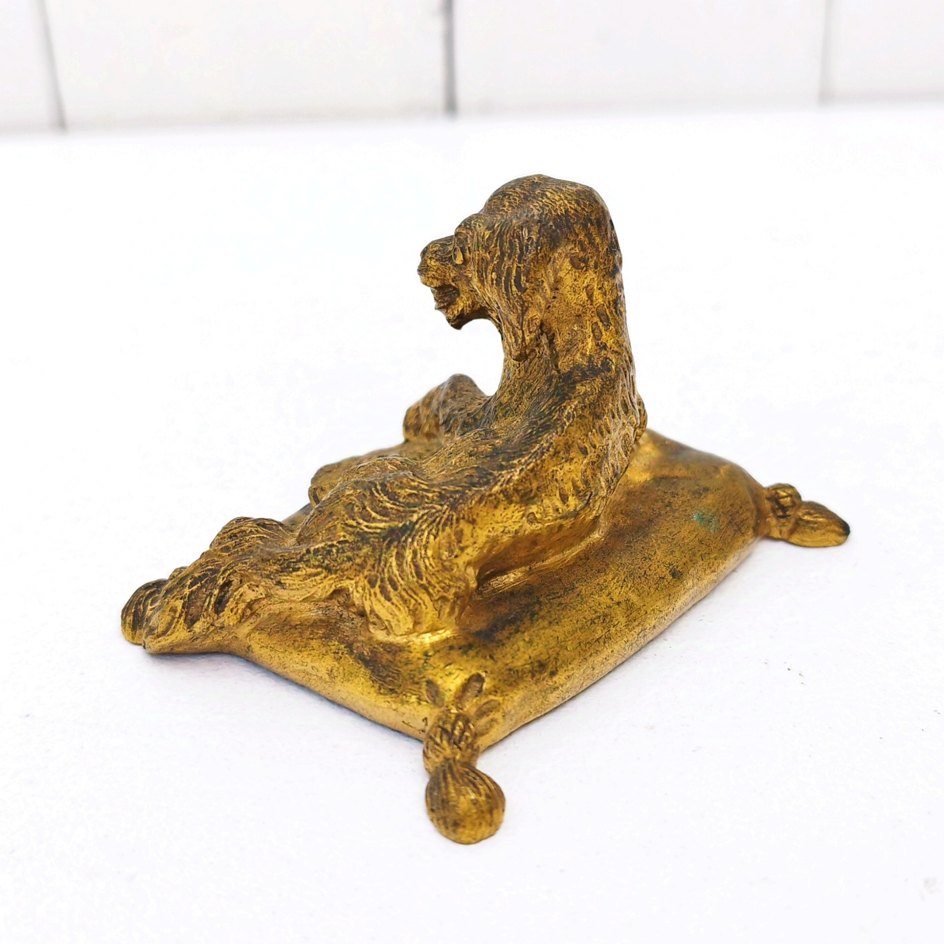 Petit bronze doré représentant un chien assis sur un coussin. quatre pieds représentant les pompons du coussin. Dorure usée par endroit. Bon état général. Dimensions :  7,5 x 5 x 5 cm