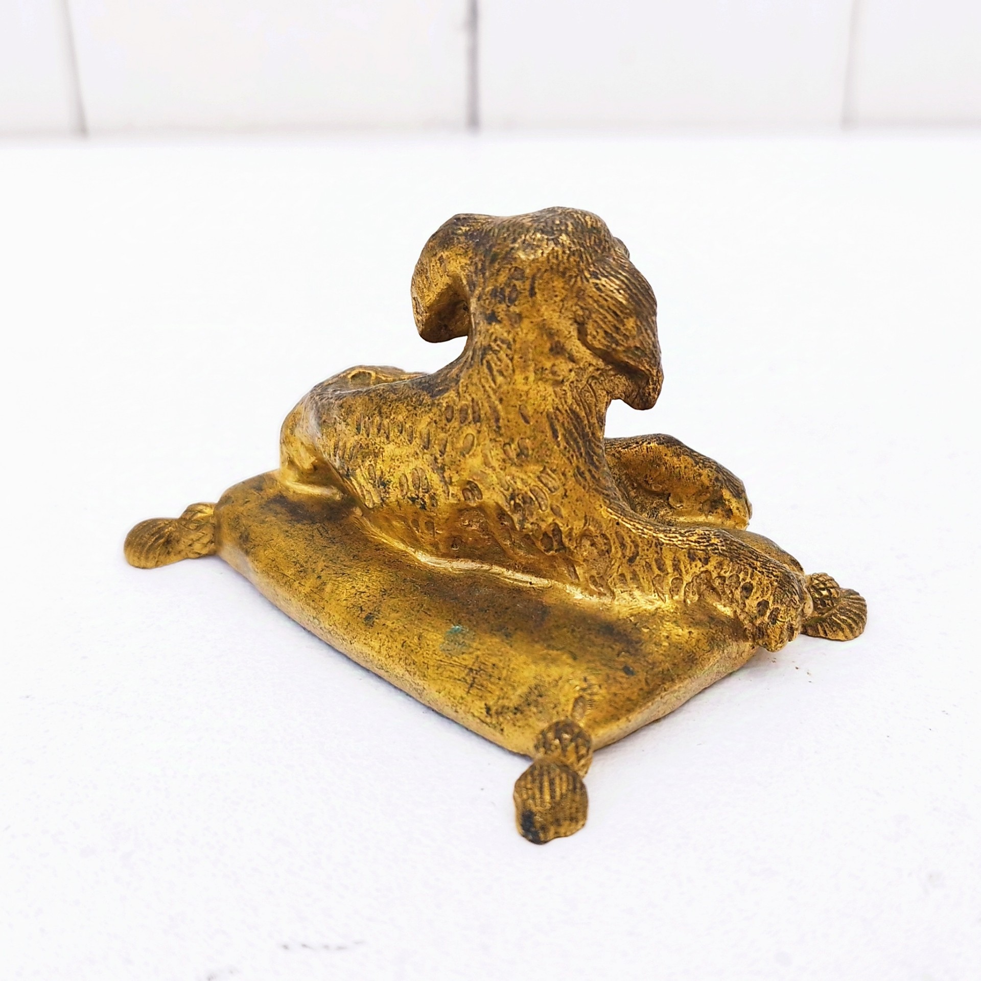 Petit bronze doré représentant un chien assis sur un coussin. quatre pieds représentant les pompons du coussin. Dorure usée par endroit. Bon état général. Dimensions :  7,5 x 5 x 5 cm