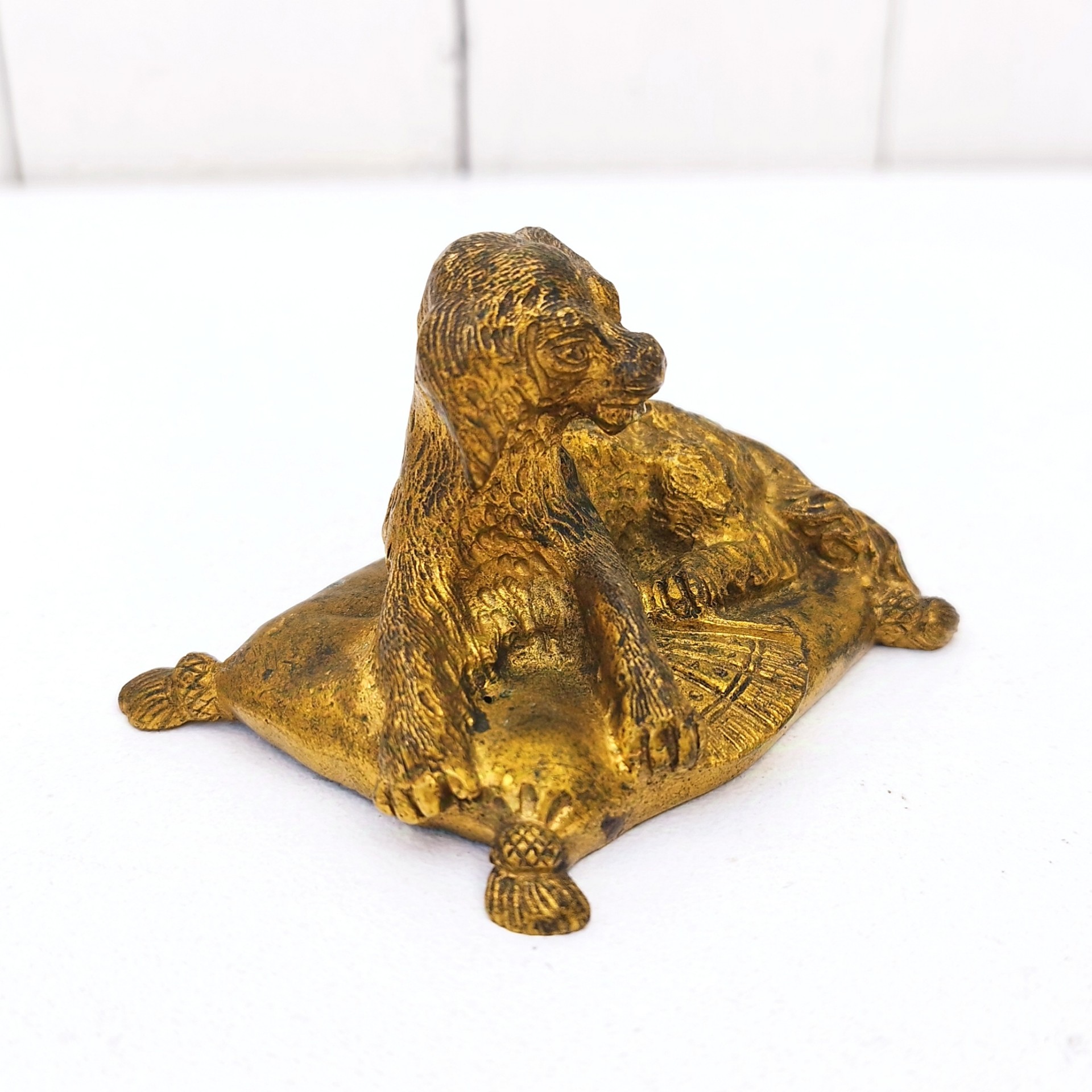 Petit bronze doré représentant un chien assis sur un coussin. quatre pieds représentant les pompons du coussin. Dorure usée par endroit. Bon état général. Dimensions :  7,5 x 5 x 5 cm
