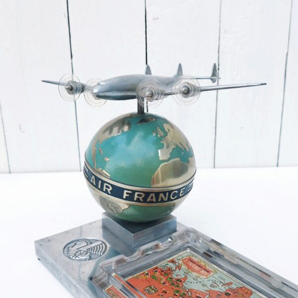 Rare authentique cendrier prestige Air France globe avec Constellation en fonte d'aluminium peinte, vers 1950. Représentant un Lockheed Constellation survolant un globe terrestre avec un cendrier en verre décoré d'un mappemonde par Lucien Boucher.  Globe cerclé du message " Air France - Réseau aérien mondial" Socle décoré du logo Air France hippocampe moulé dans la masse, globe peint (peinture jaunie sur la partie basse du globe, à cause de la fumée de cigarette) .  Constellation avec hélices à double rayures. Marqué Made in France sur le socle à l'arrière du globe. Décor de la mappemonde du cendrier écaillé sur le contour, traces d'usage sur le socle, patins en caoutchouc noir tous présents et en bon état. Ensemble en très bon état d'origine. Hauteur : ~17 cm Longueur : 24,4 cm Profondeur : 12,8 cm Constellation : 21 x 17 cm