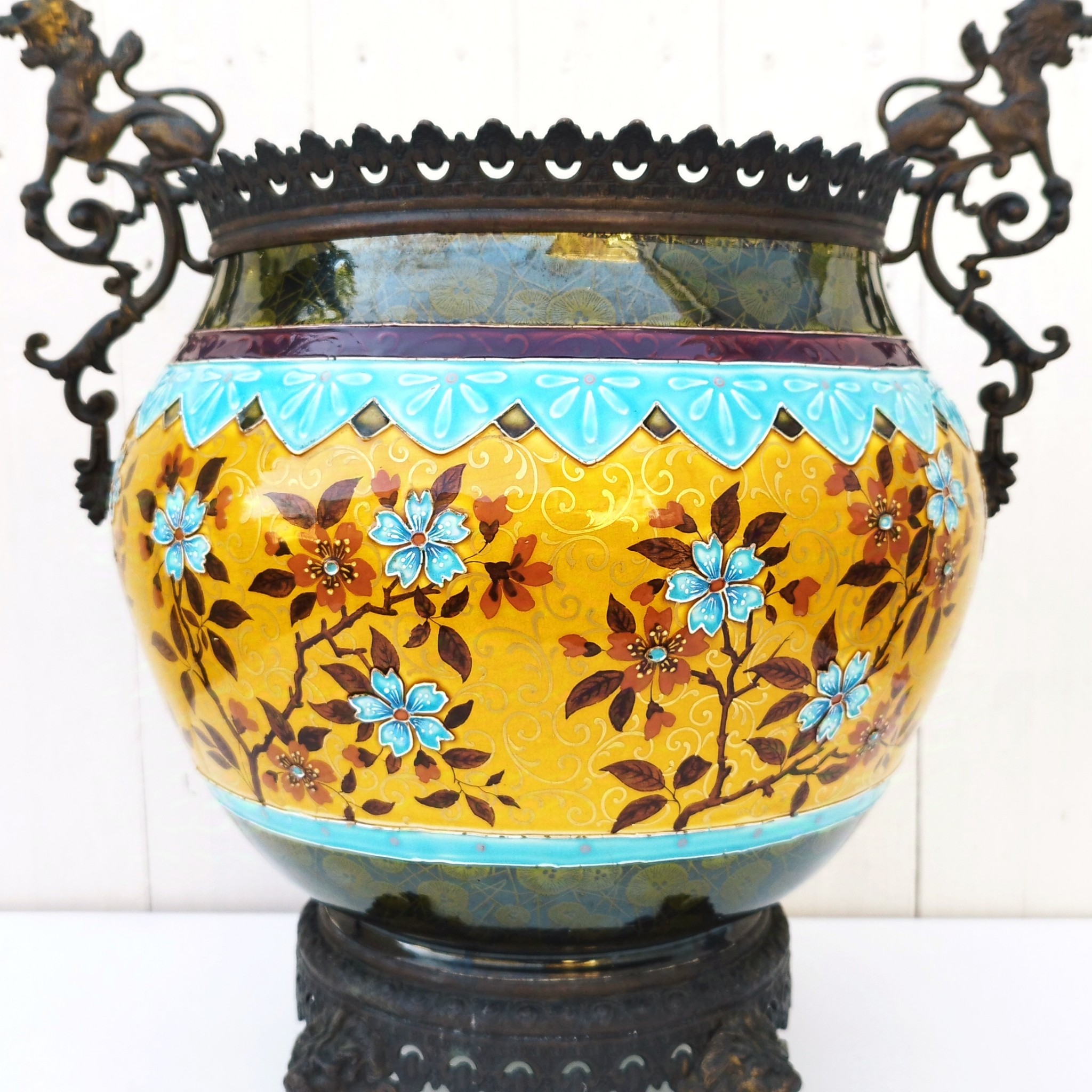 Grand cache-pot ou jardinière en céramique de Sarreguemines numéro 2048. Cerclé d'une couronne en bronze aux prises formant des lions. Pied tripodes en bronze. Couleurs bien vives. races d'usage à l'intérieur. Très bon état. Hauteur max  : 40 cm Diamètre : 25 cm