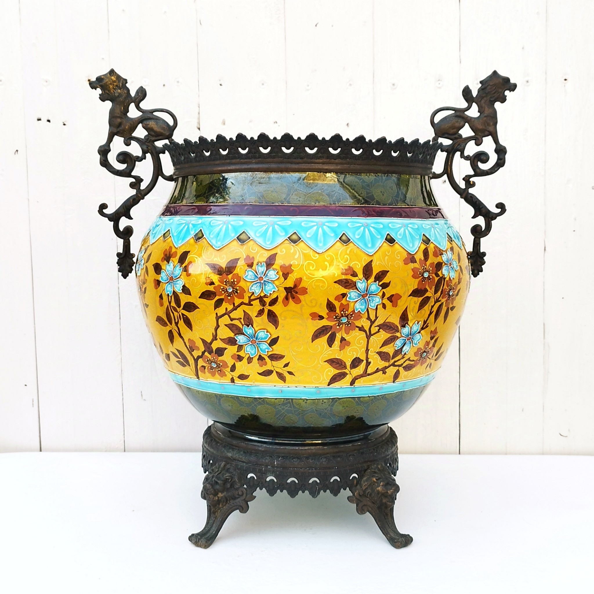 Grand cache-pot ou jardinière en céramique de Sarreguemines numéro 2048. Cerclé d'une couronne en bronze aux prises formant des lions. Pied tripodes en bronze. Couleurs bien vives. races d'usage à l'intérieur. Très bon état. Hauteur max  : 40 cm Diamètre : 25 cm