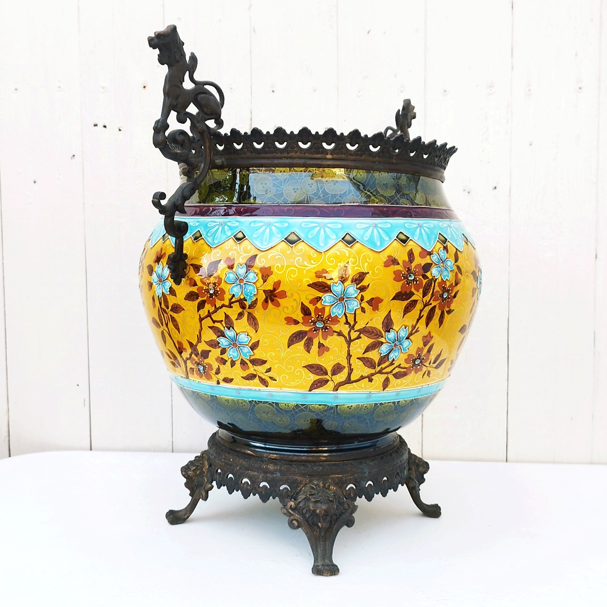 Grand cache-pot ou jardinière en céramique de Sarreguemines numéro 2048. Cerclé d'une couronne en bronze aux prises formant des lions. Pied tripodes en bronze. Couleurs bien vives. races d'usage à l'intérieur. Très bon état. Hauteur max  : 40 cm Diamètre : 25 cm