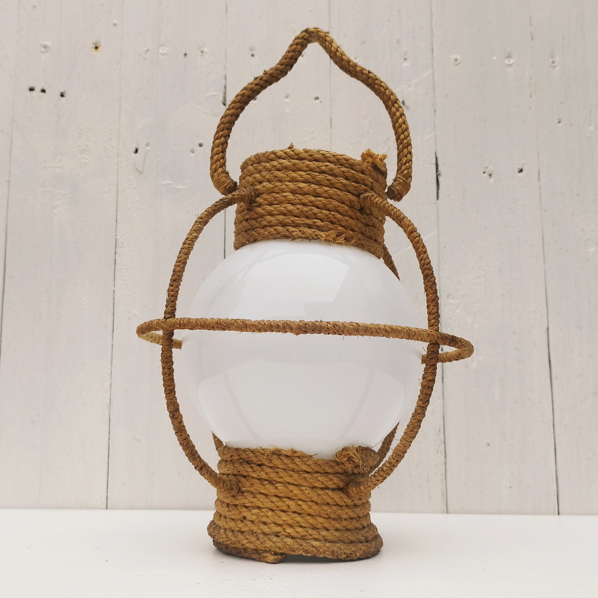 Suspension datant des années 50, à la forme d'une lanterne/lampe tempête créée par Audoux Minet. Structure en fil d'acier et métal recouvert de cordage tressé. Globe en opaline blanche. Manque sa douille à l'intérieur. Petit décollement de cordage sur le haut. Bon état général. Hauteur  avec anse: 36 cm