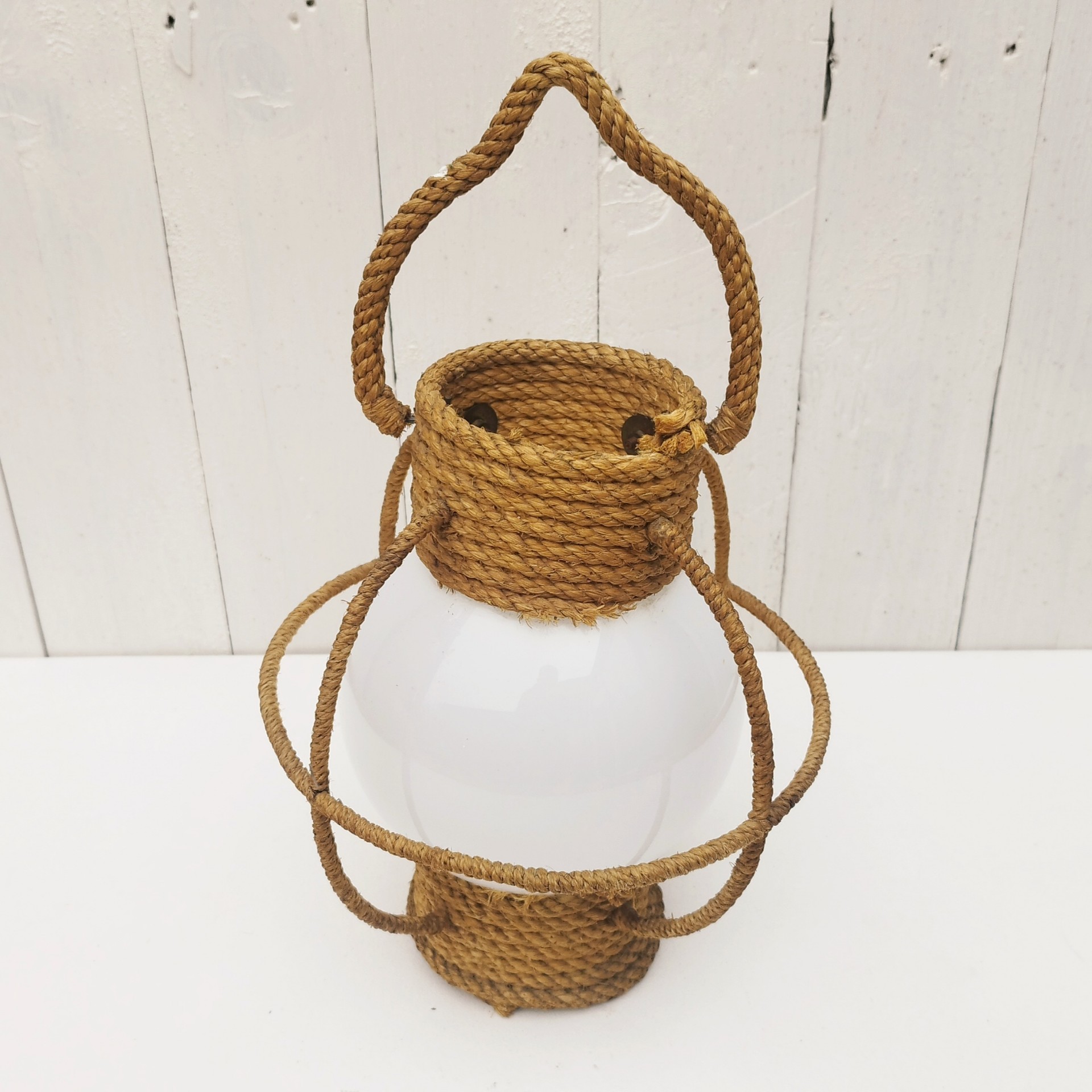 Suspension datant des années 50, à la forme d'une lanterne/lampe tempête créée par Audoux Minet. Structure en fil d'acier et métal recouvert de cordage tressé. Globe en opaline blanche. Manque sa douille à l'intérieur. Petit décollement de cordage sur le haut. Bon état général. Hauteur  avec anse: 36 cm