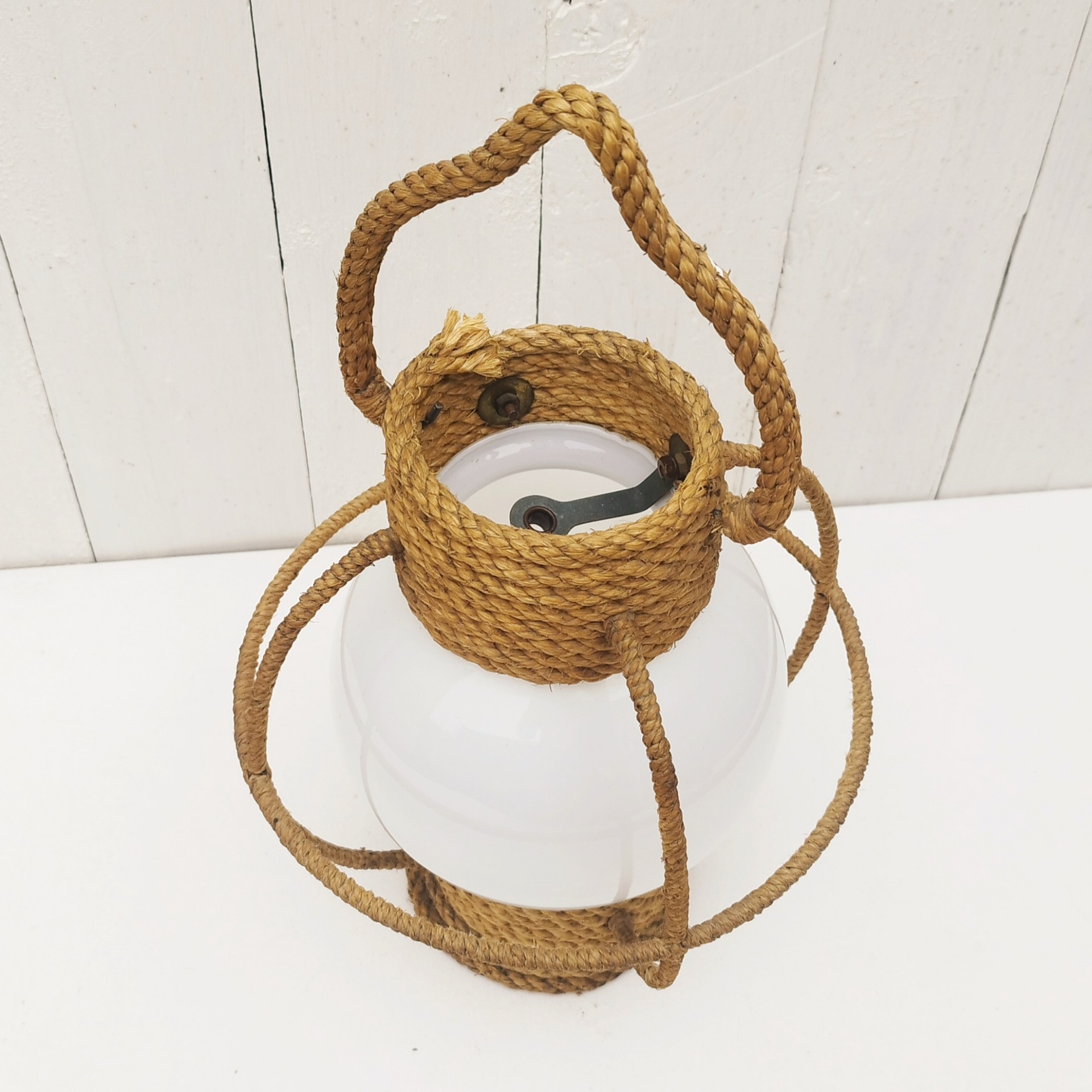 Suspension datant des années 50, à la forme d'une lanterne/lampe tempête créée par Audoux Minet. Structure en fil d'acier et métal recouvert de cordage tressé. Globe en opaline blanche. Manque sa douille à l'intérieur. Petit décollement de cordage sur le haut. Bon état général. Hauteur  avec anse: 36 cm