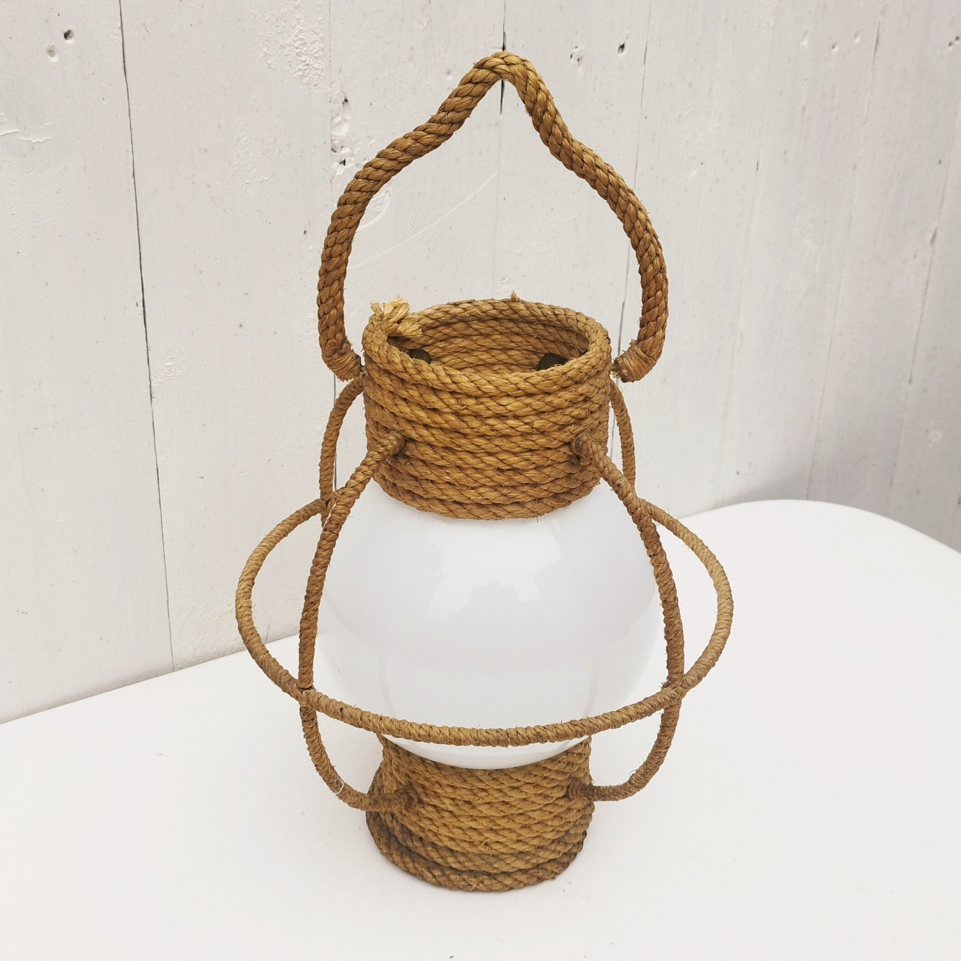Suspension datant des années 50, à la forme d'une lanterne/lampe tempête créée par Audoux Minet. Structure en fil d'acier et métal recouvert de cordage tressé. Globe en opaline blanche. Manque sa douille à l'intérieur. Petit décollement de cordage sur le haut. Bon état général. Hauteur  avec anse: 36 cm