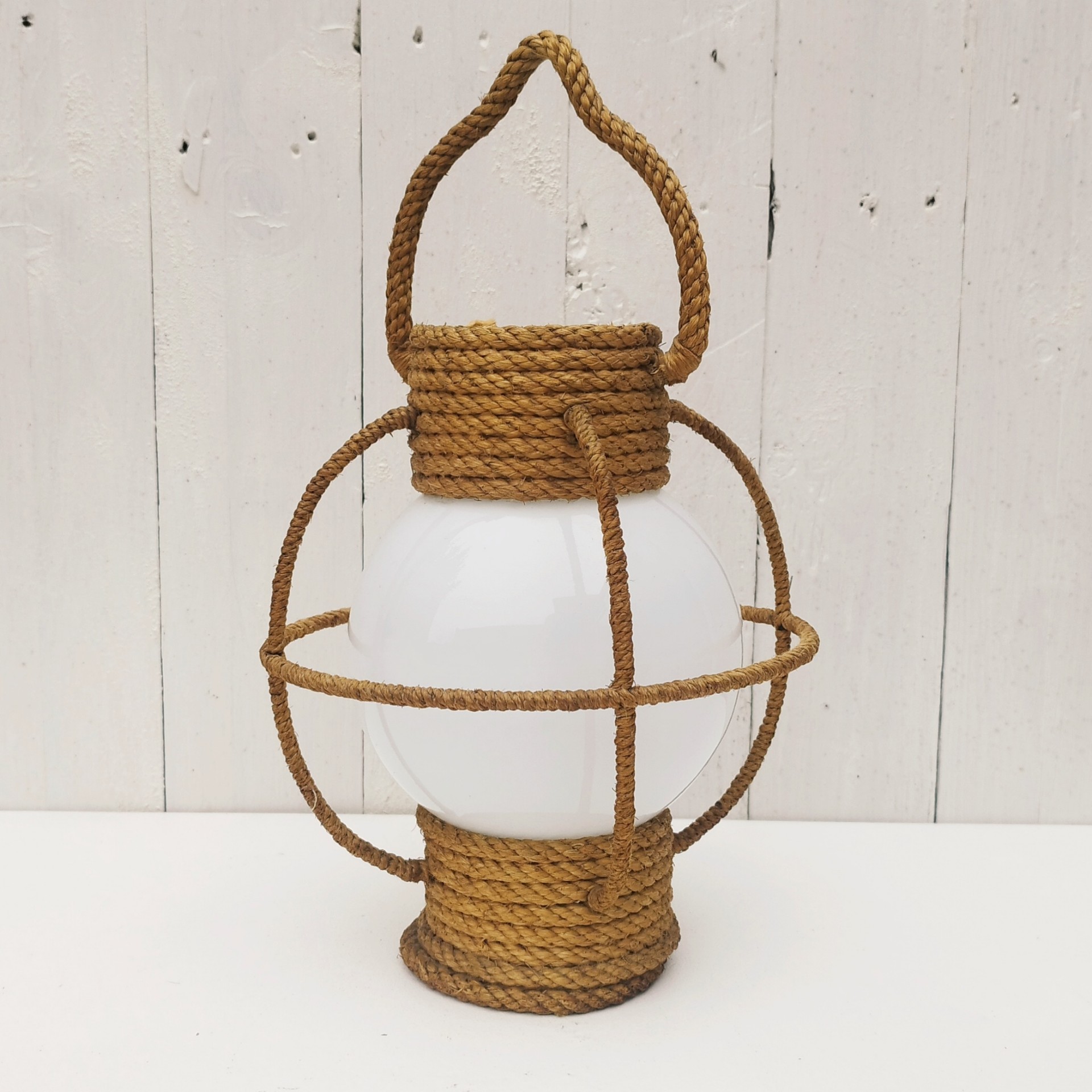Suspension datant des années 50, à la forme d'une lanterne/lampe tempête créée par Audoux Minet. Structure en fil d'acier et métal recouvert de cordage tressé. Globe en opaline blanche. Manque sa douille à l'intérieur. Petit décollement de cordage sur le haut. Bon état général. Hauteur  avec anse: 36 cm