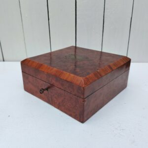 Boite à bijoux en bois  vernis  avec écusson en laiton eu centre du couvercle. Capitonnage en satin bleu. rayures et petites taches d'usage. Un choc sur l'arête droite. Possède sa clé. Bon état général Dimensions : 19 x 19 cm Hauteur fermée : 8 cm