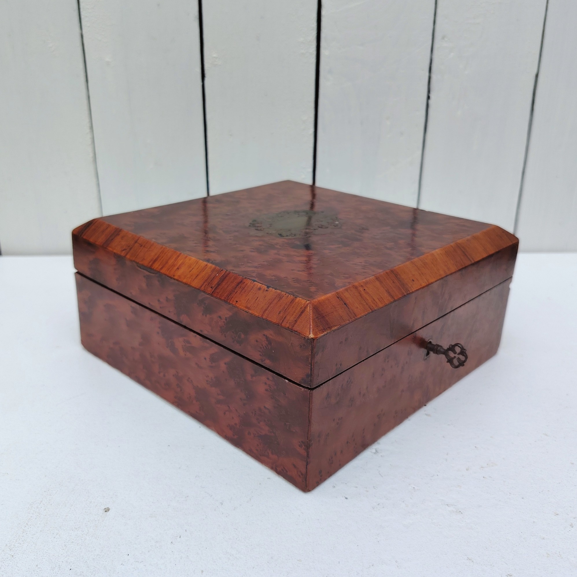 Boite à bijoux en bois  vernis  avec écusson en laiton eu centre du couvercle. Capitonnage en satin bleu. rayures et petites taches d'usage. Un choc sur l'arête droite. Possède sa clé. Bon état général Dimensions : 19 x 19 cm Hauteur fermée : 8 cm