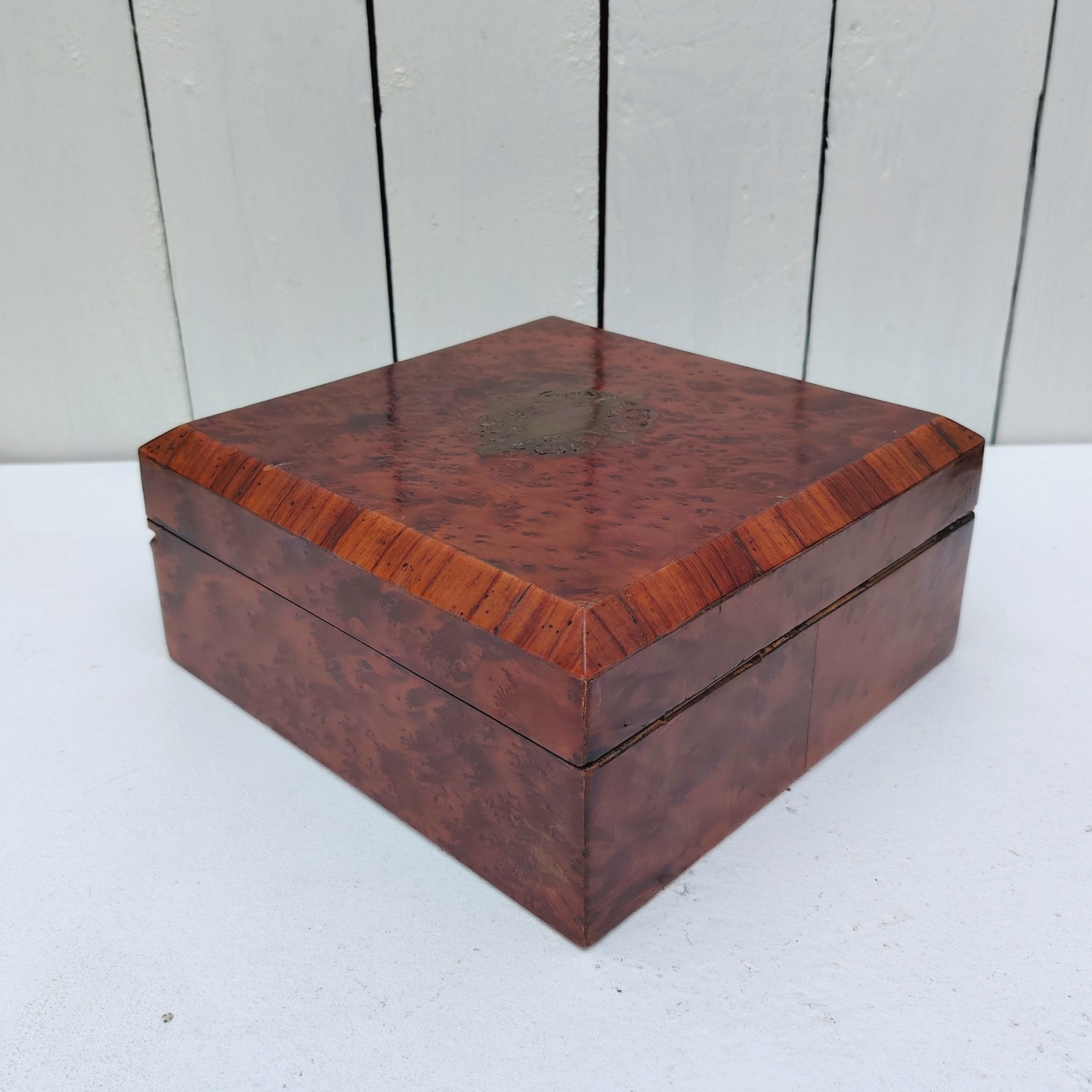 Boite à bijoux en bois  vernis  avec écusson en laiton eu centre du couvercle. Capitonnage en satin bleu. rayures et petites taches d'usage. Un choc sur l'arête droite. Possède sa clé. Bon état général Dimensions : 19 x 19 cm Hauteur fermée : 8 cm