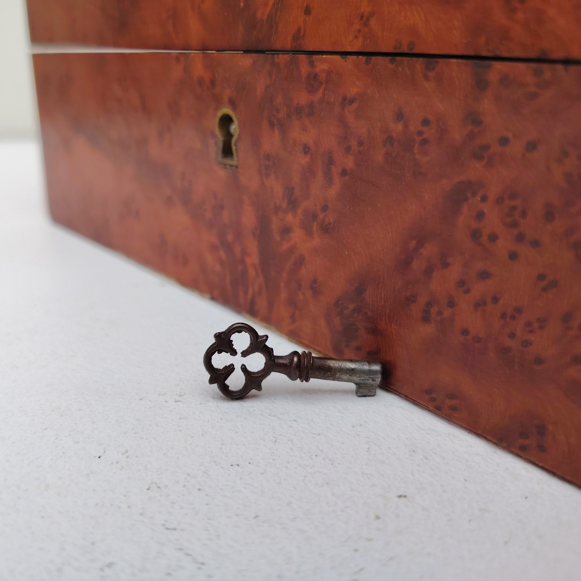 Boite à bijoux en bois  vernis  avec écusson en laiton eu centre du couvercle. Capitonnage en satin bleu. rayures et petites taches d'usage. Un choc sur l'arête droite. Possède sa clé. Bon état général Dimensions : 19 x 19 cm Hauteur fermée : 8 cm