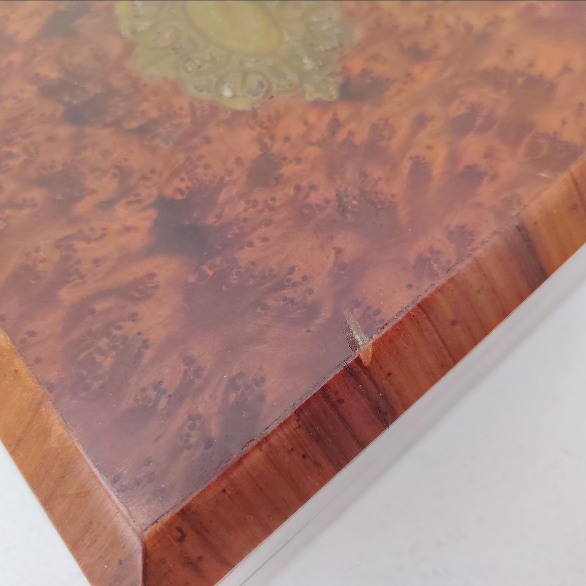 Boite à bijoux en bois  vernis  avec écusson en laiton eu centre du couvercle. Capitonnage en satin bleu. rayures et petites taches d'usage. Un choc sur l'arête droite. Possède sa clé. Bon état général Dimensions : 19 x 19 cm Hauteur fermée : 8 cm