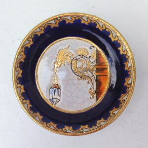 Vide poche, coupelle ou ramasse pièce en faïence de Longwy, à décor d'une lanterne  au bout d'une enseigne, émaux dorés. Certainement une commande spéciale d'un magasin ou hotel. Effet craquelé sur la céramique. Signée sur le dessous. Une micro égrenure sur le contour. Très bon état. Diamètre : 13,5 cm Hauteur : 3 cm