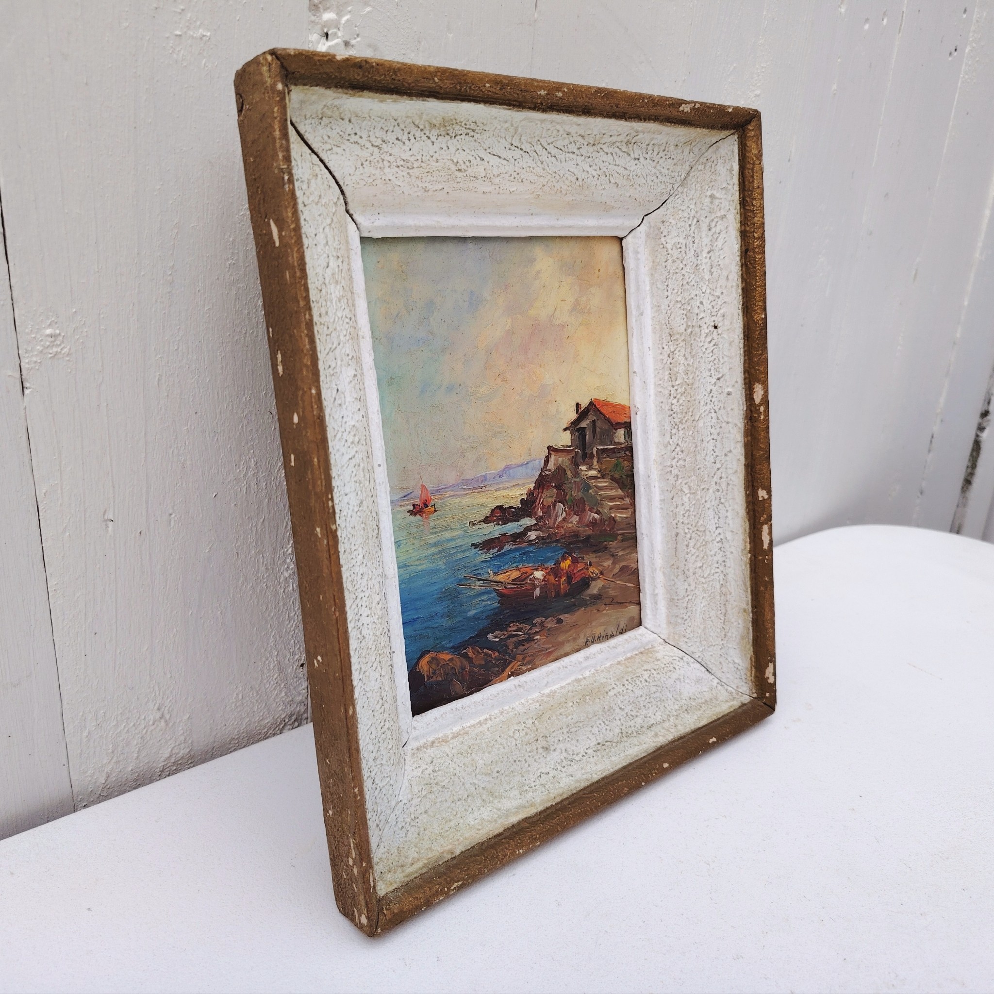 Huile sur panneau isorel représentant un bateau amarré dans une crique en premier plan , en deuxième plan une maison de pêcheur sur une hauteur et dans le fond on distingue une falaise et un voilier naviguant. Cadre en bois cérusé blanc. Signé RU Rinaldi en bas à droite. Bon état général. Dimensions cadre : 22 x 25,5 cm