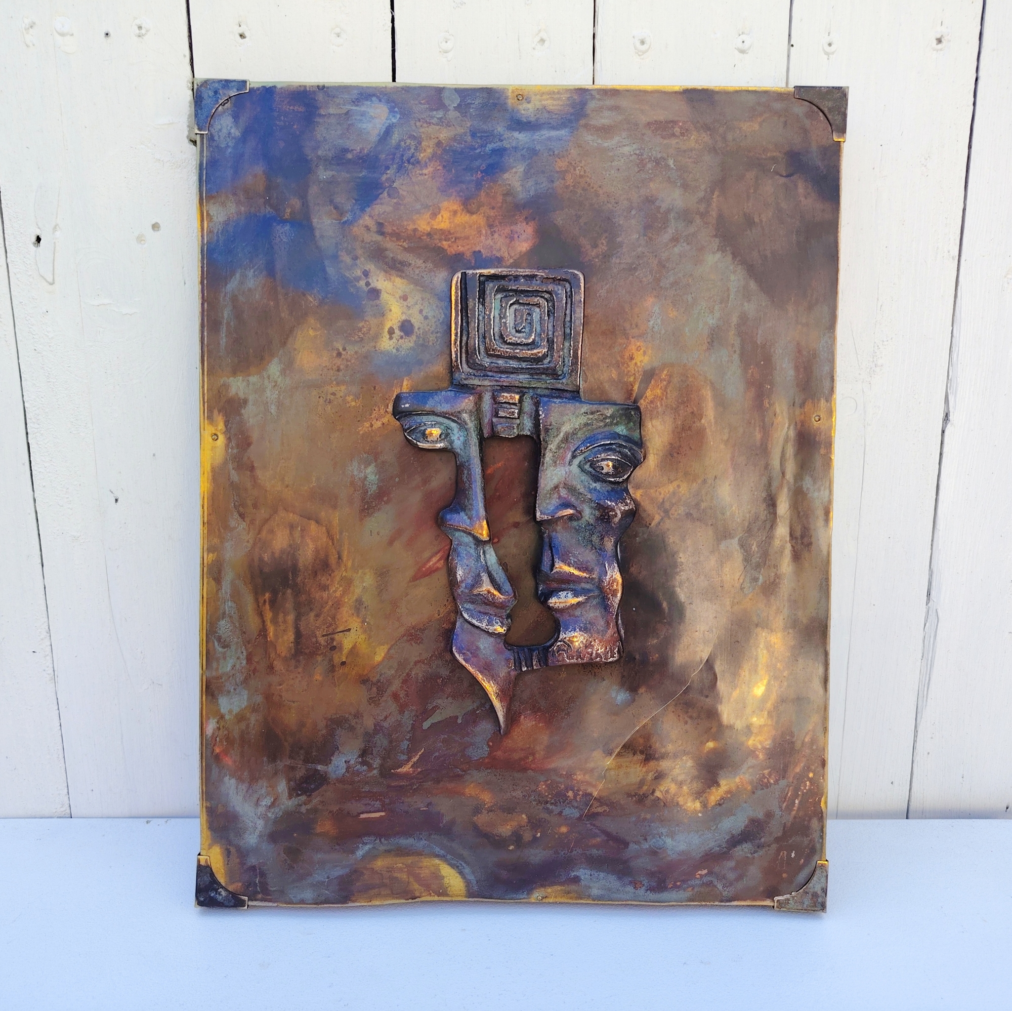 Plaque en cuivre avec deux visages en reliefs au centre qui sont en métal patiné bronze, le tout collé sur un panneau de bois très épais. Signé en bas d'un visage. Bon état général. Dimensions : 38,5 x 30 cm