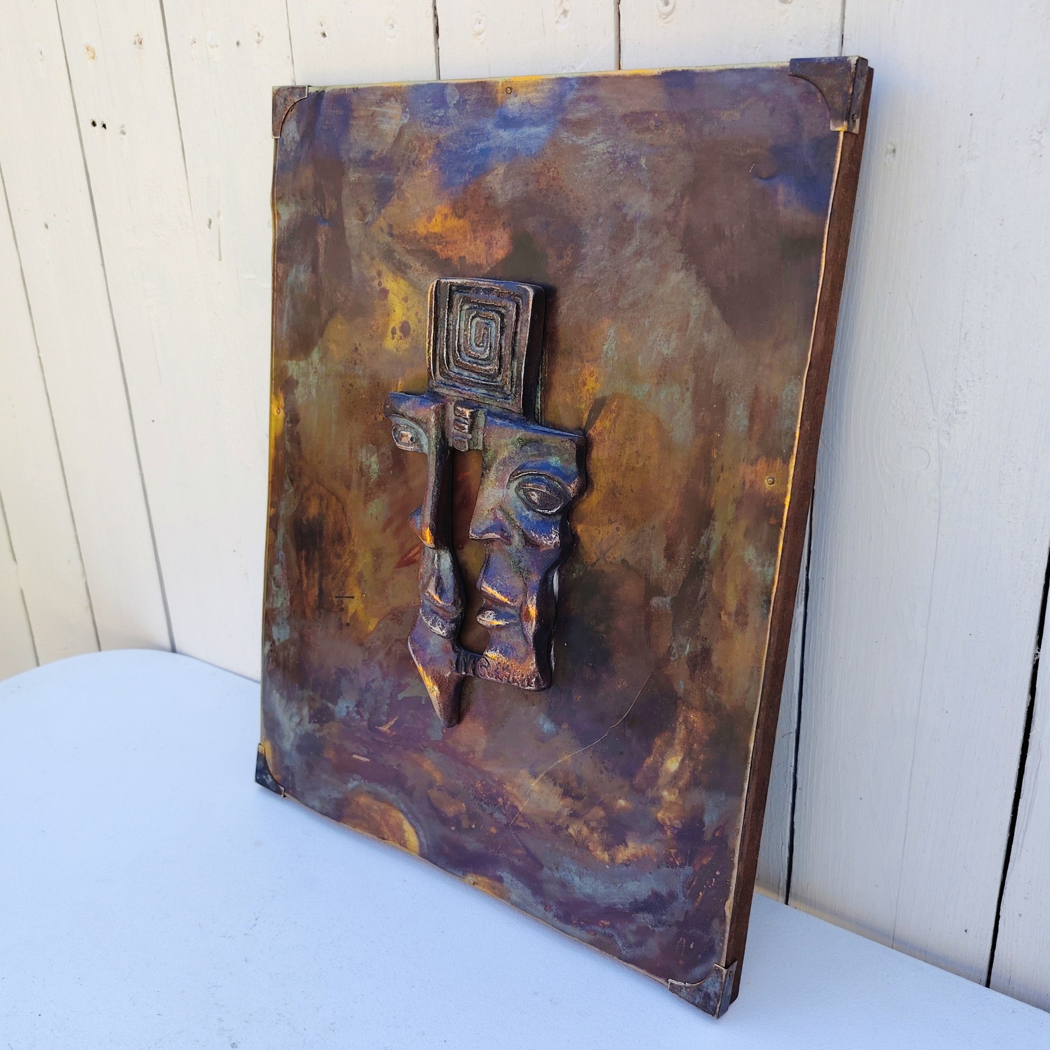Plaque en cuivre avec deux visages en reliefs au centre qui sont en métal patiné bronze, le tout collé sur un panneau de bois très épais. Signé en bas d'un visage. Bon état général. Dimensions : 38,5 x 30 cm