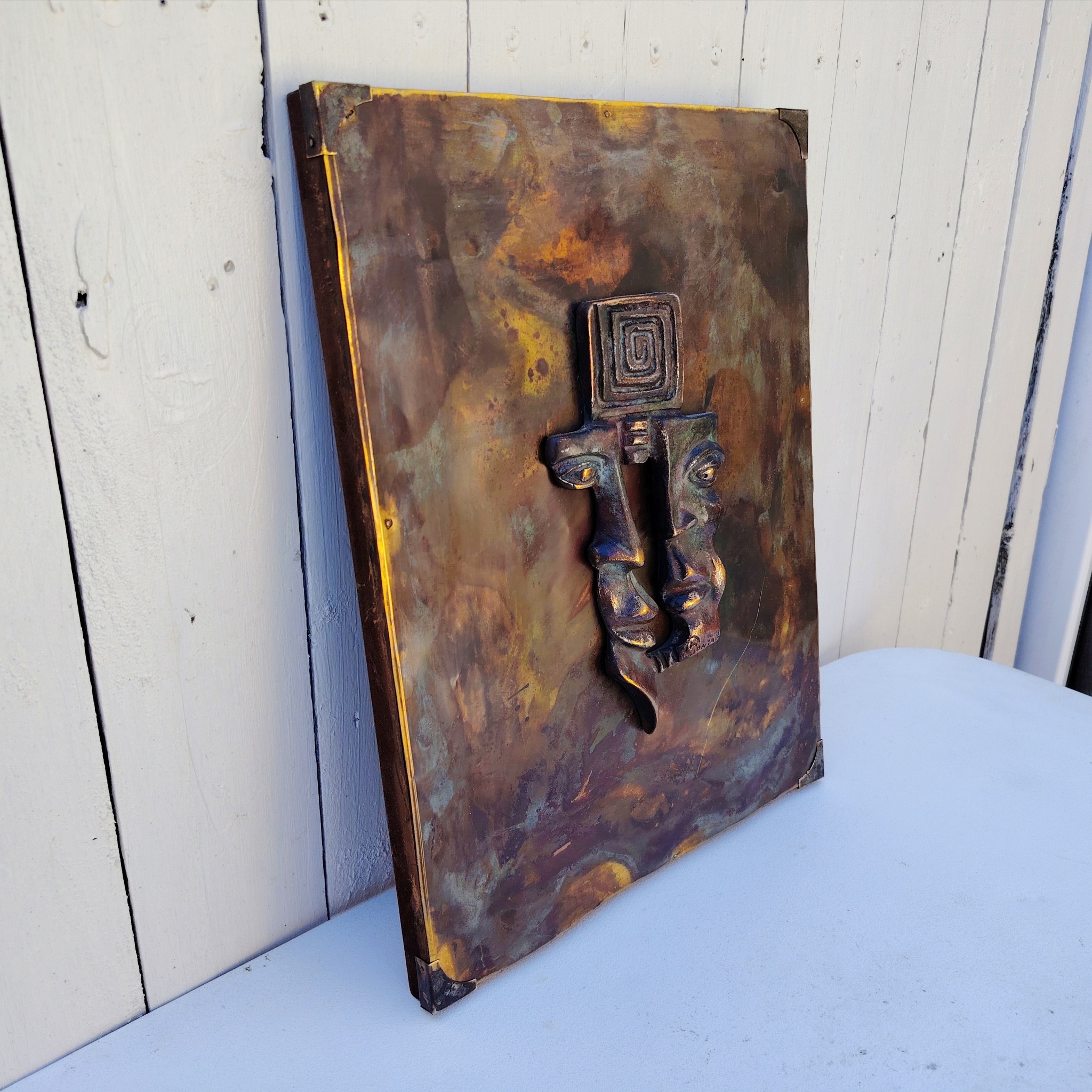 Plaque en cuivre avec deux visages en reliefs au centre qui sont en métal patiné bronze, le tout collé sur un panneau de bois très épais. Signé en bas d'un visage. Bon état général. Dimensions : 38,5 x 30 cm