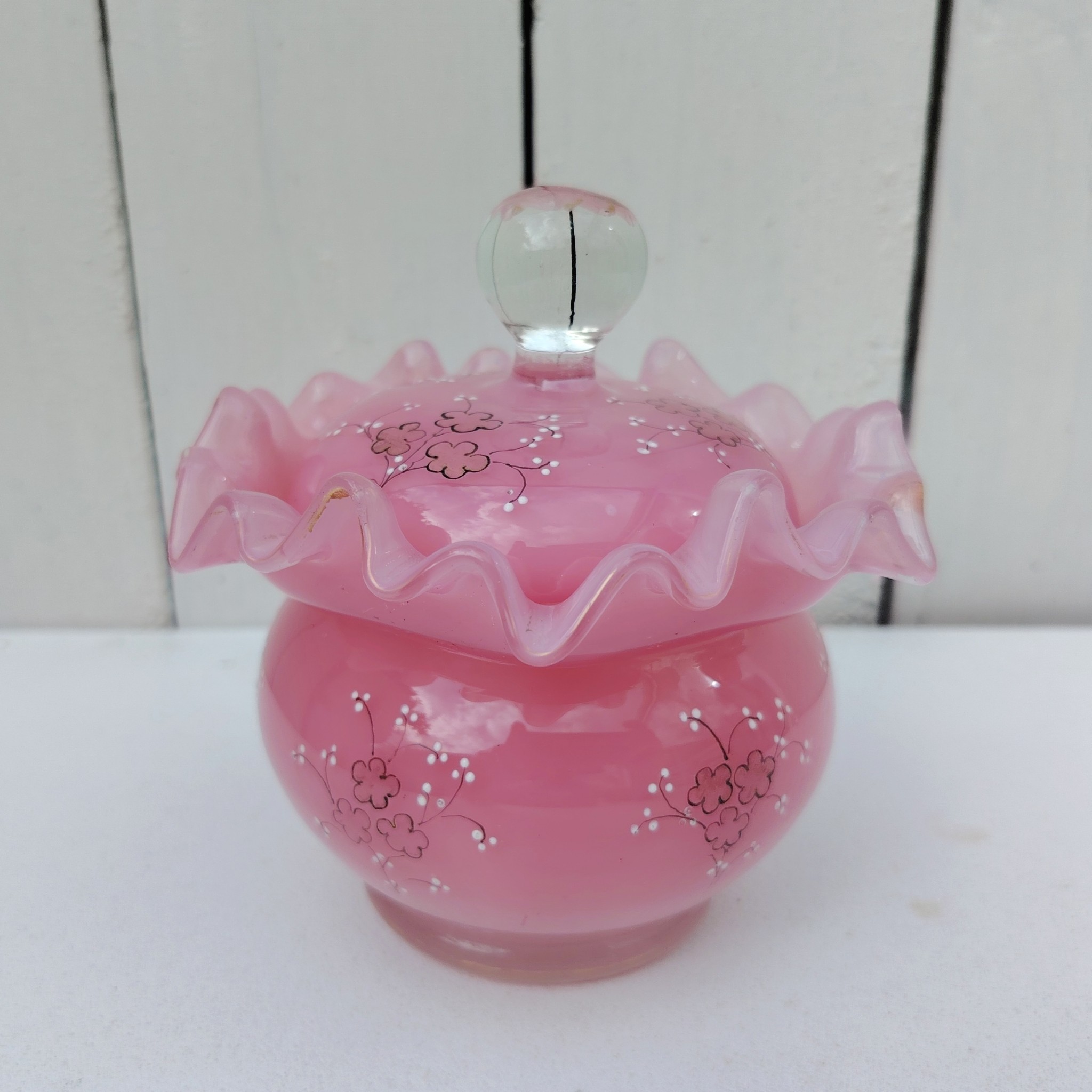 Pot couvert pouvant servir de baguier ou autre en verre soufflé de couleur rose, collerette torsadée. Décor de fleur rehaussé de points émaillés blancs en relief. Les fleurs et la prise du couvercle devait être certainement doré car on y voit encore le petites de la dorure. Un petit éclat sur l'intérieur du couvercle non visible quand il est fermé, et traces blanches sur le talon du pot non visible quand il est posé. Bon état général Hauteur avec couvercle : 13 cm Diamètre : 11 cm