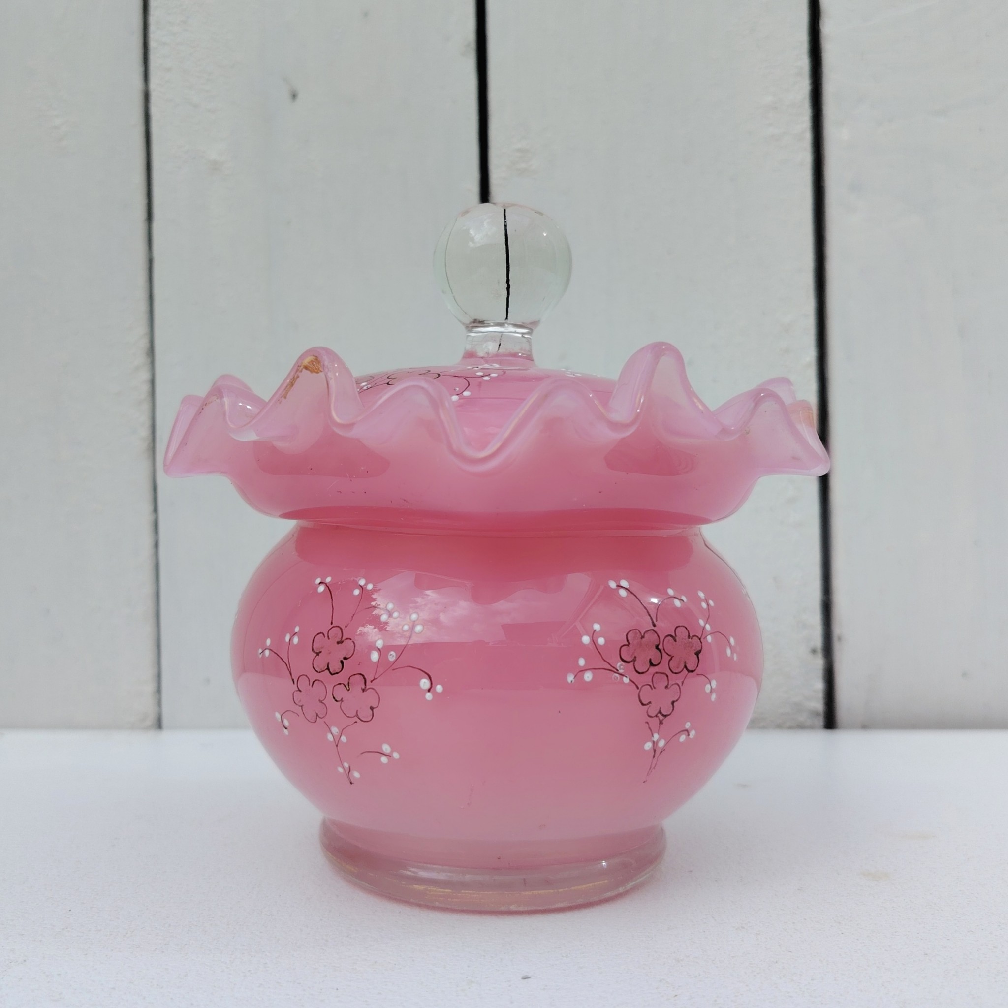 Pot couvert pouvant servir de baguier ou autre en verre soufflé de couleur rose, collerette torsadée. Décor de fleur rehaussé de points émaillés blancs en relief. Les fleurs et la prise du couvercle devait être certainement doré car on y voit encore le petites de la dorure. Un petit éclat sur l'intérieur du couvercle non visible quand il est fermé, et traces blanches sur le talon du pot non visible quand il est posé. Bon état général Hauteur avec couvercle : 13 cm Diamètre : 11 cm
