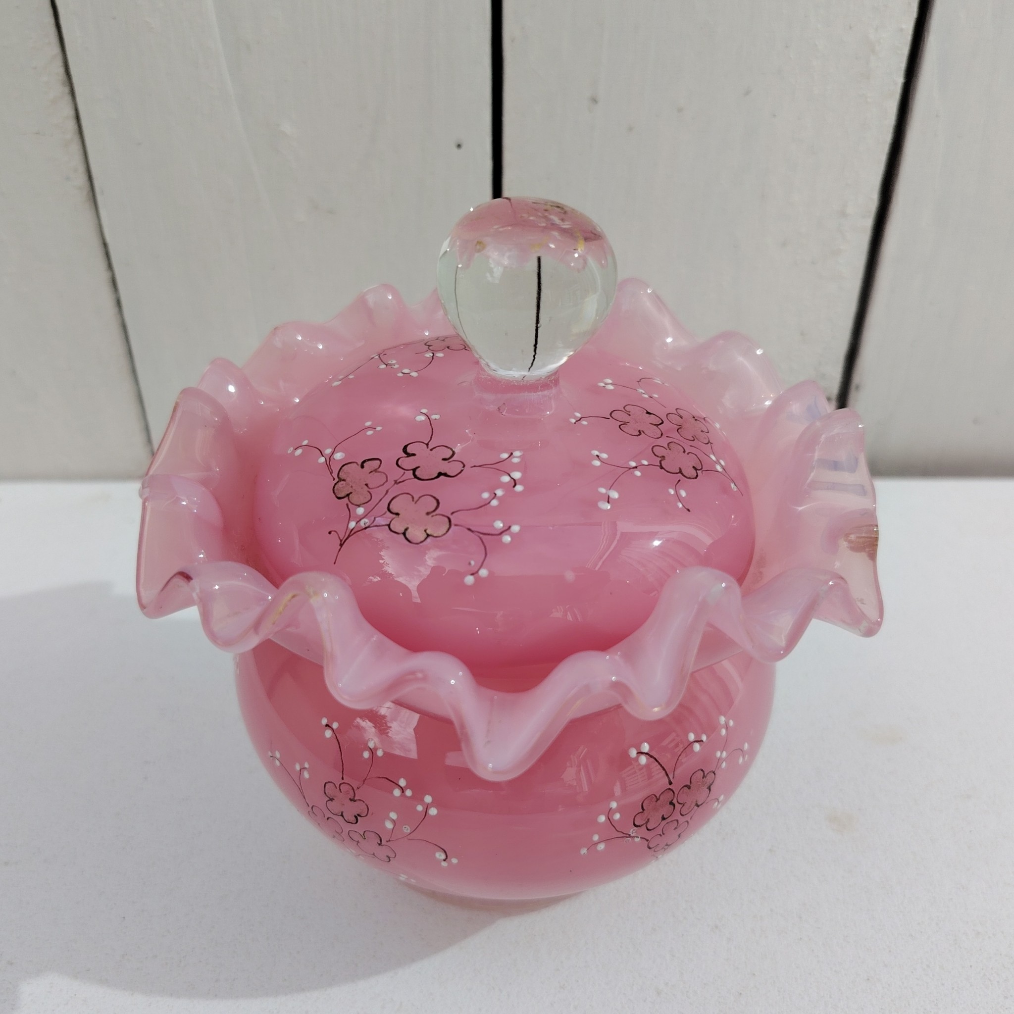 Pot couvert pouvant servir de baguier ou autre en verre soufflé de couleur rose, collerette torsadée. Décor de fleur rehaussé de points émaillés blancs en relief. Les fleurs et la prise du couvercle devait être certainement doré car on y voit encore le petites de la dorure. Un petit éclat sur l'intérieur du couvercle non visible quand il est fermé, et traces blanches sur le talon du pot non visible quand il est posé. Bon état général Hauteur avec couvercle : 13 cm Diamètre : 11 cm