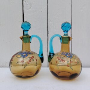 Paire de carafes Georges Sand en verre de couleur ocre et anse bleue turquoise, décor floral émaillé. Petits éclats sur l'intérieur du col d'une carafe non visible quand le bouchon y est  placé. Manque de dorure sur les bouchons et les anses Bon état général Hauteur avec bouchon : 17 cm