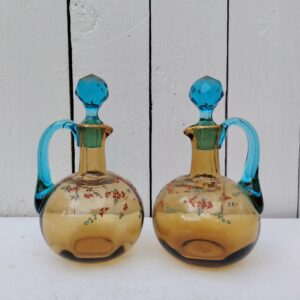 Paire de carafes Georges Sand en verre de couleur ocre et anse bleue turquoise, décor floral émaillé. Petits éclats sur l'intérieur du col d'une carafe non visible quand le bouchon y est  placé. Manque de dorure sur les bouchons et les anses Bon état général Hauteur avec bouchon : 17 cm