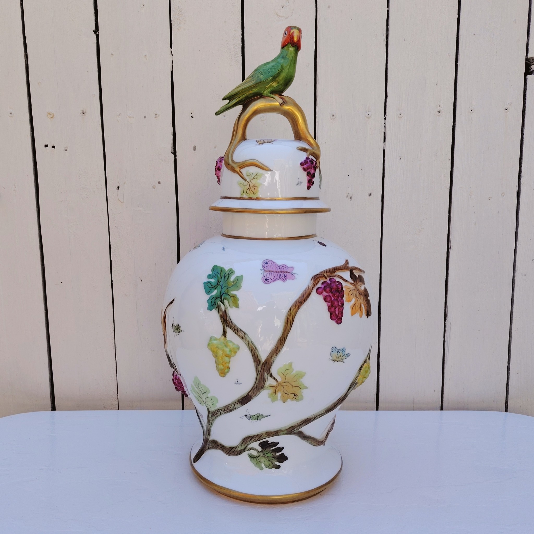 Grand pot couvert en porcelaine portugaise Pac. Orné de vignes et grappes de raisins en relief, et de papillons et coccinelles peints. Liseré doré, poignée du couvercle représentant une branche dorée surhaussé d'un perroquet. Très belle facture. Excellent état.  Hauteur avec couvercle : ~47 cm