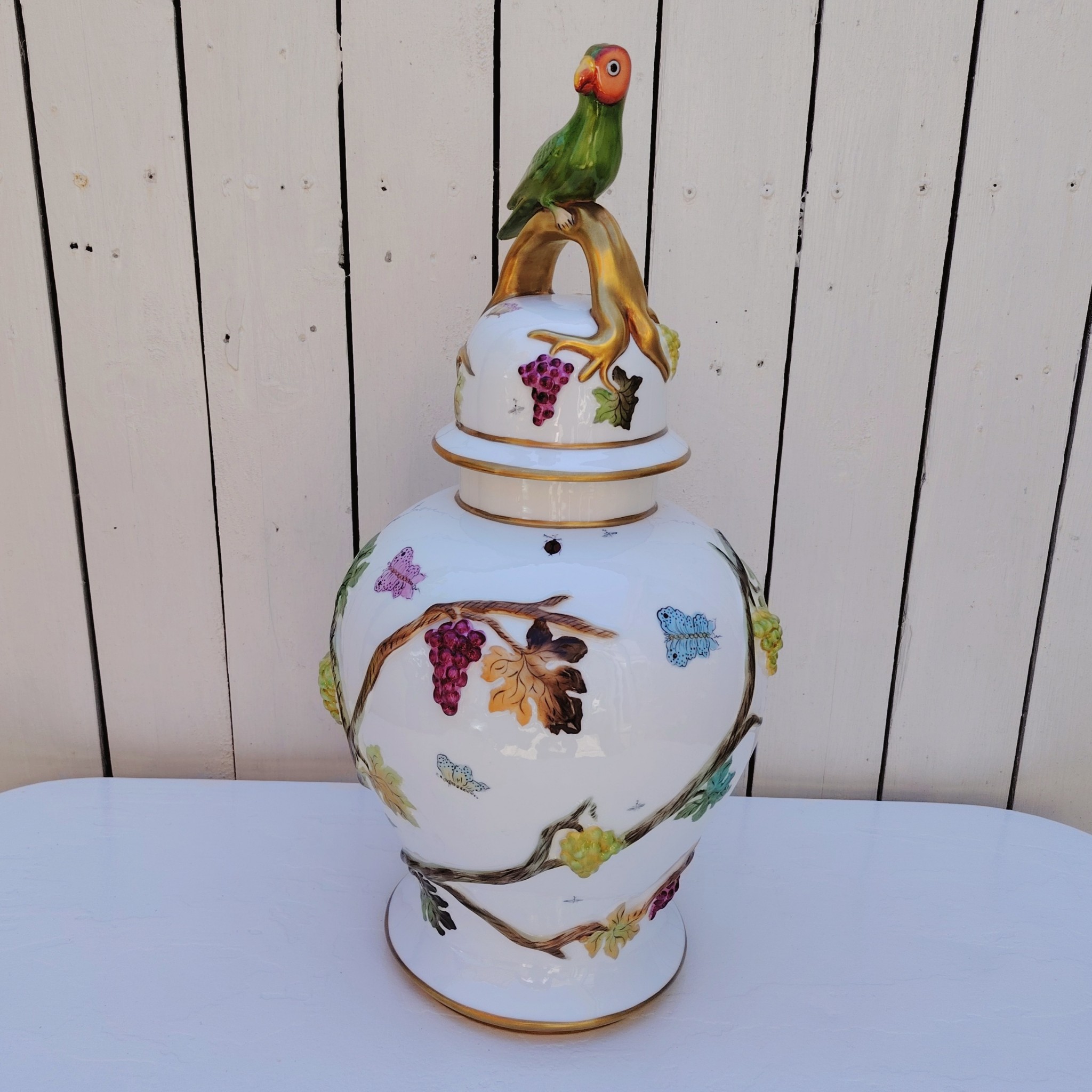 Grand pot couvert en porcelaine portugaise Pac. Orné de vignes et grappes de raisins en relief, et de papillons et coccinelles peints. Liseré doré, poignée du couvercle représentant une branche dorée surhaussé d'un perroquet. Très belle facture. Excellent état.  Hauteur avec couvercle : ~47 cm