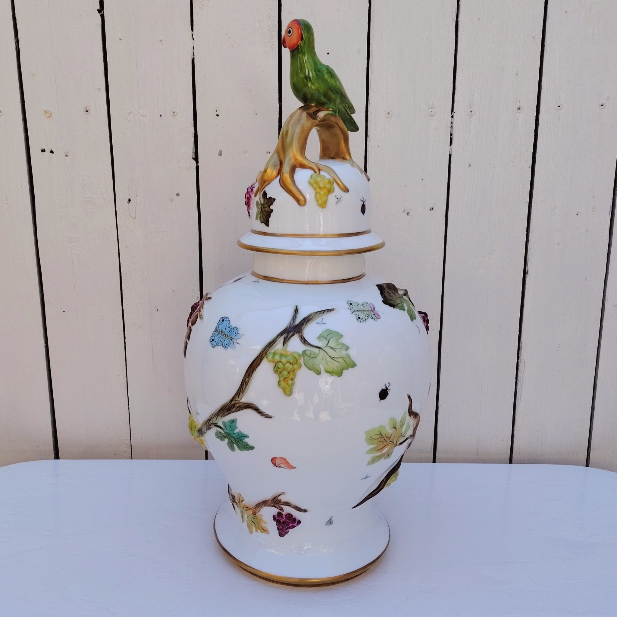 Grand pot couvert en porcelaine portugaise Pac. Orné de vignes et grappes de raisins en relief, et de papillons et coccinelles peints. Liseré doré, poignée du couvercle représentant une branche dorée surhaussé d'un perroquet. Très belle facture. Excellent état.  Hauteur avec couvercle : ~47 cm