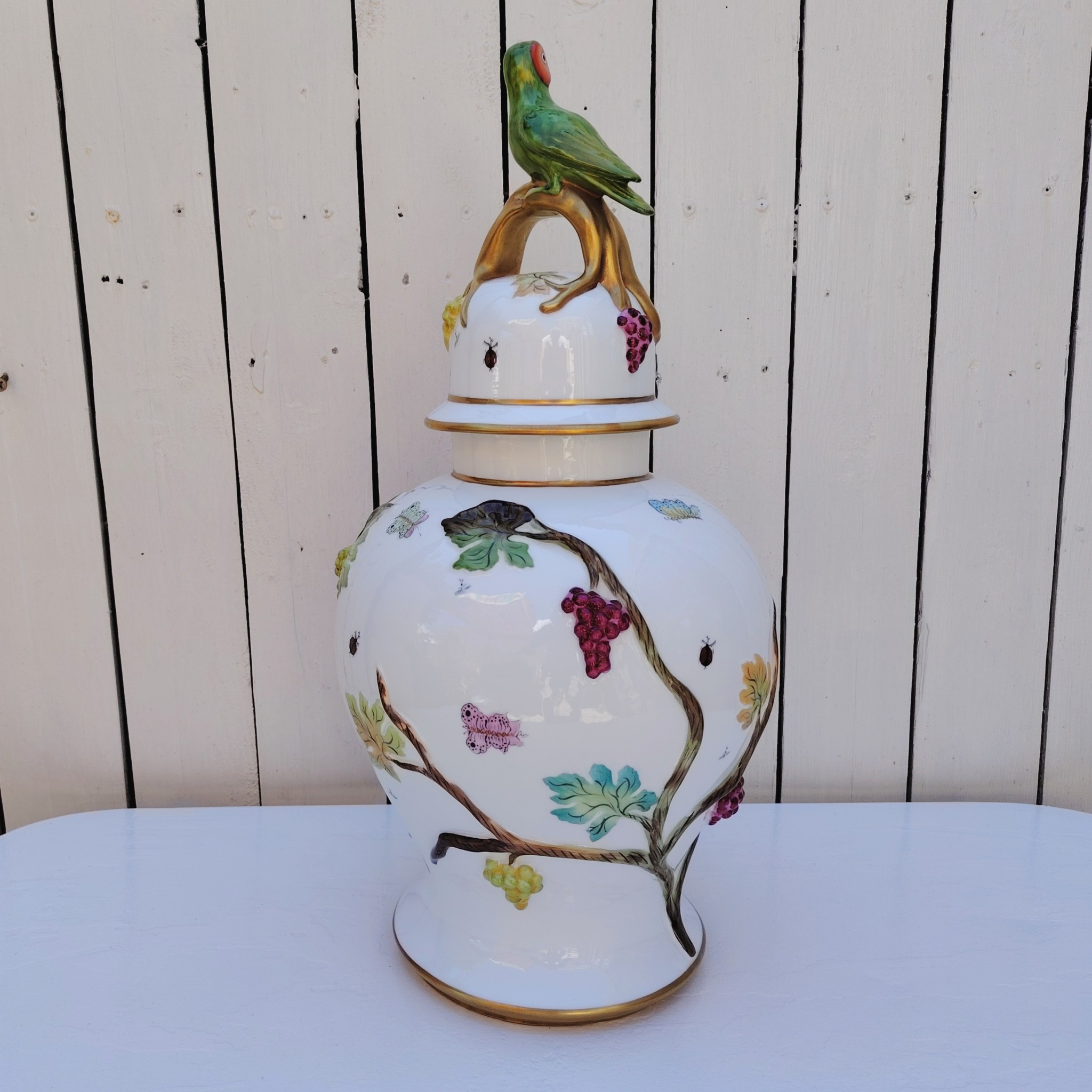 Grand pot couvert en porcelaine portugaise Pac. Orné de vignes et grappes de raisins en relief, et de papillons et coccinelles peints. Liseré doré, poignée du couvercle représentant une branche dorée surhaussé d'un perroquet. Très belle facture. Excellent état.  Hauteur avec couvercle : ~47 cm