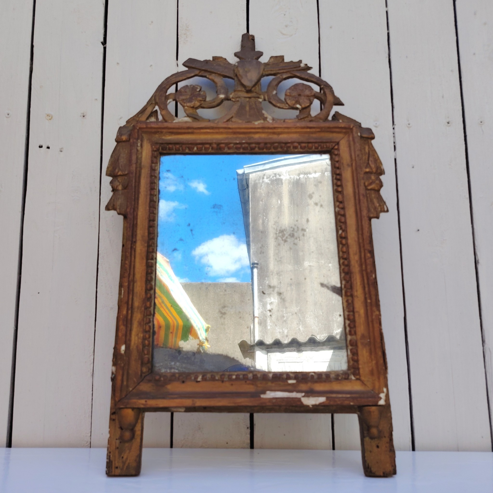Ancien miroir en bois et stuc doré, de style Louis XVI, fin XVIIIéme début XIXéme. Fronton à enroulement de feuillage orné d'un coeur en son centre. Manque une partie du fronton, des éclats et maque de dorure. Tain terni et taché. Dans son jus Dimensions : 54 x 33 x 4 cm