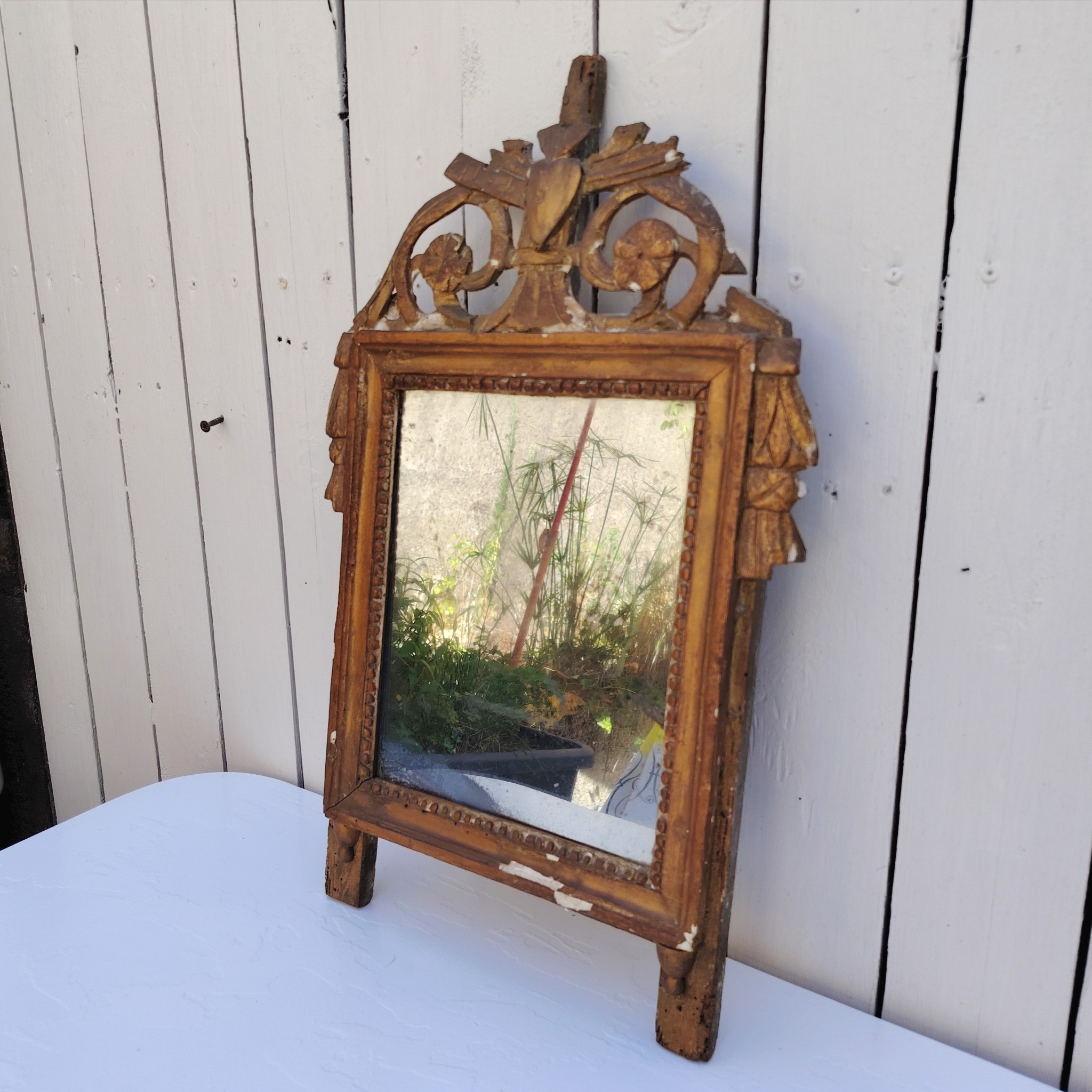Ancien miroir en bois et stuc doré, de style Louis XVI, fin XVIIIéme début XIXéme. Fronton à enroulement de feuillage orné d'un coeur en son centre. Manque une partie du fronton, des éclats et maque de dorure. Tain terni et taché. Dans son jus Dimensions : 54 x 33 x 4 cm