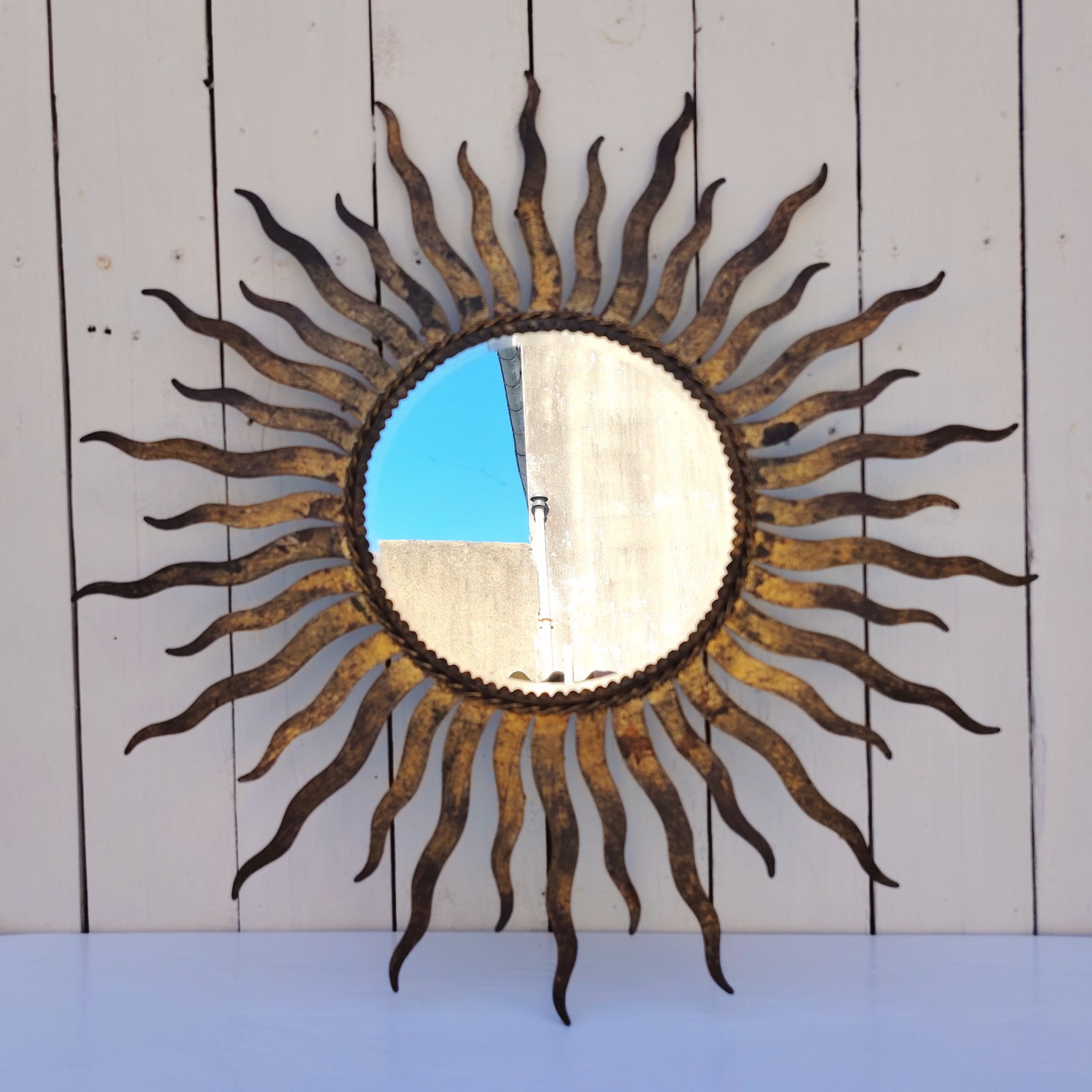 Miroir soleil, oeil de sorcière, en métal patiné doré datant des années 50. Miroir biseauté. Traces de corrosion et manques de dorures. Un point d'accroche sur l'arrière. Bon état général Diamètre total : 53,5 cm Diamètre miroir : 20,5 cm