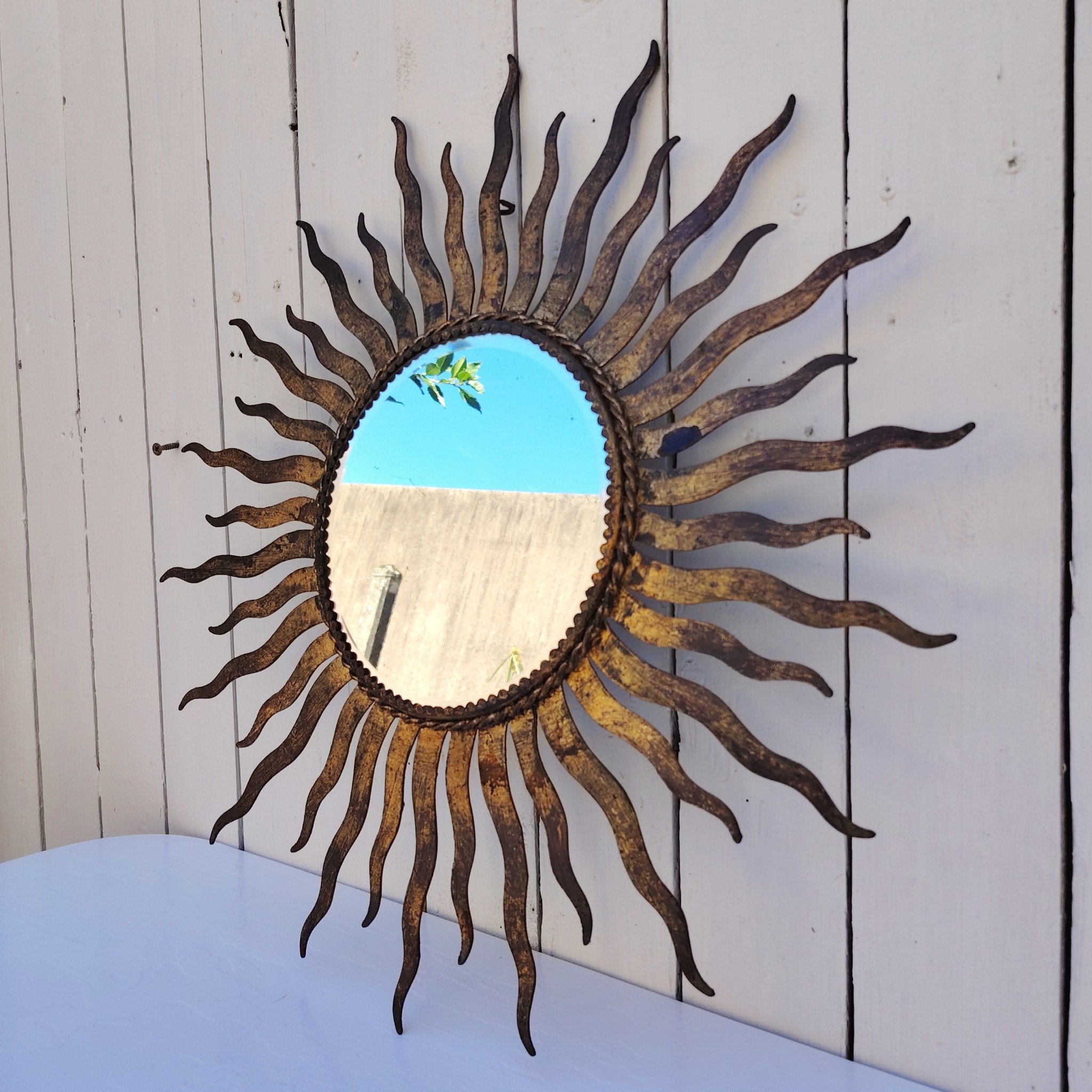 Miroir soleil, oeil de sorcière, en métal patiné doré datant des années 50. Miroir biseauté. Traces de corrosion et manques de dorures. Un point d'accroche sur l'arrière. Bon état général Diamètre total : 53,5 cm Diamètre miroir : 20,5 cm