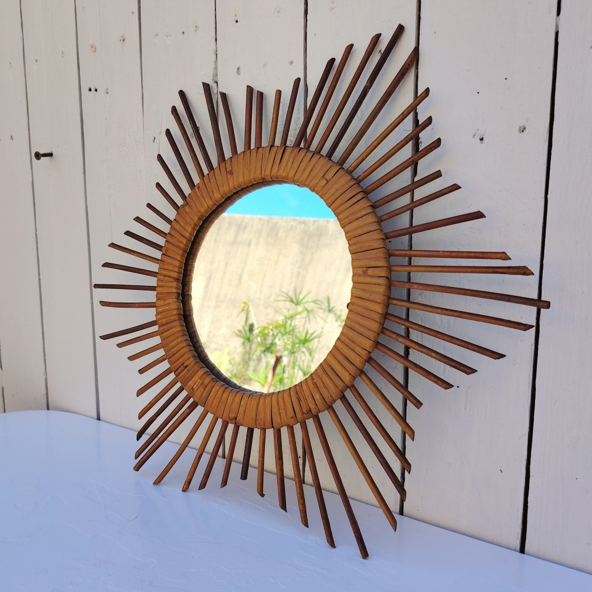  Miroir soleil en rotin datant des années 60.  Traces de  piqûres sur le miroir. Un point d'accroche sur l'arrière.  Bon état général. Diamètre total : 42  cm Diamètre miroir : 17,5 cm