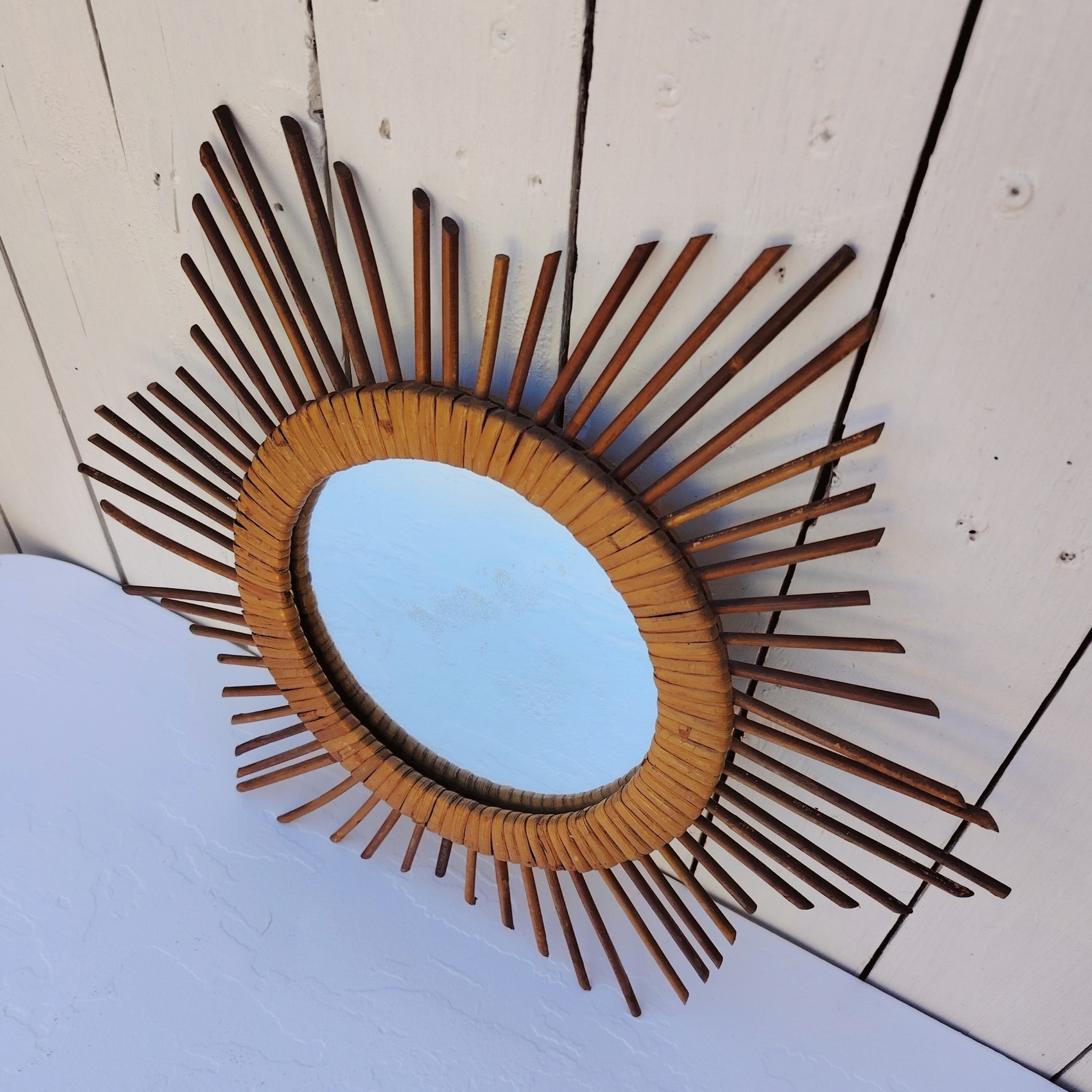  Miroir soleil en rotin datant des années 60.  Traces de  piqûres sur le miroir. Un point d'accroche sur l'arrière.  Bon état général. Diamètre total : 42  cm Diamètre miroir : 17,5 cm