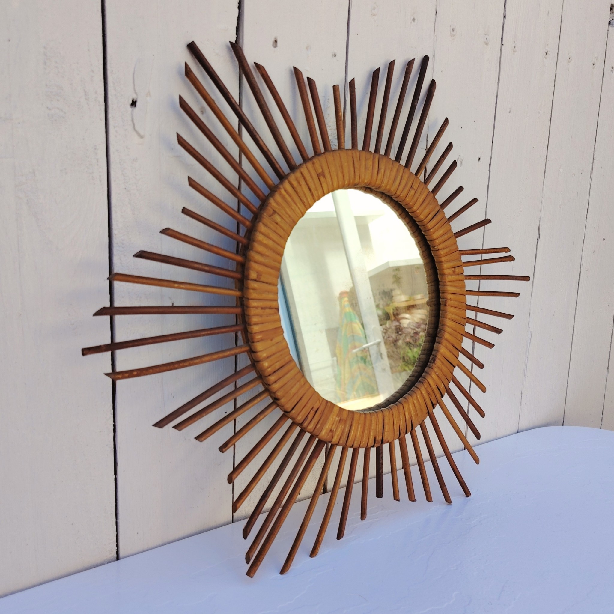  Miroir soleil en rotin datant des années 60.  Traces de  piqûres sur le miroir. Un point d'accroche sur l'arrière.  Bon état général. Diamètre total : 42  cm Diamètre miroir : 17,5 cm